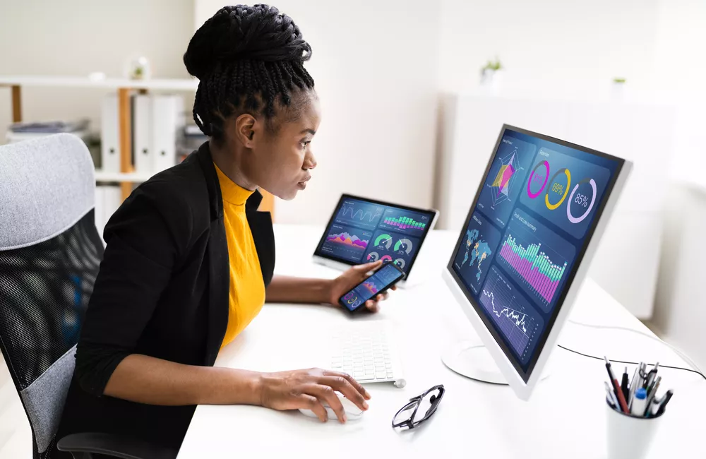 A data analyst sits at her desk analyzing data with her computer, tablet, and smartphone.