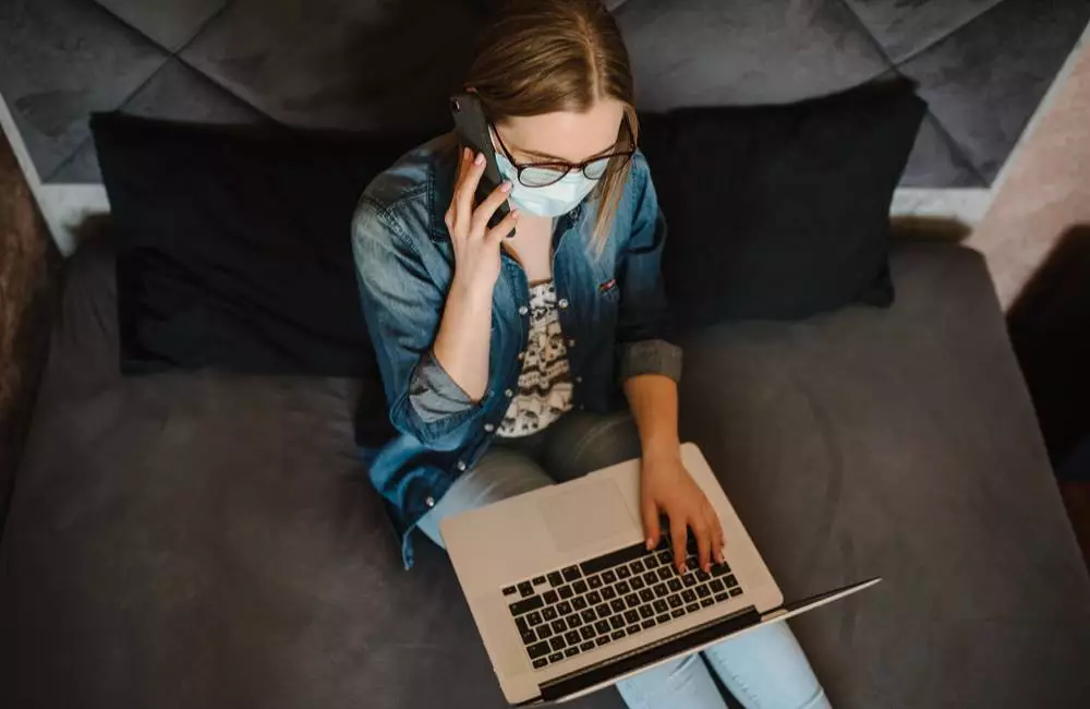 A woman wearing a mask speaks on the phone as she looks at her laptop.