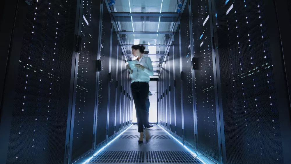 A woman holding a tablet walks down the aisle of a server room.