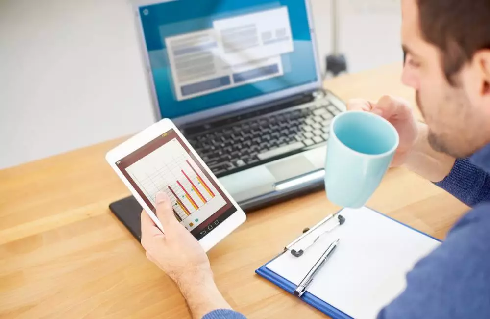 A man studies a tablet with a bar chart on it as he sits at a desk with a cup of coffee.