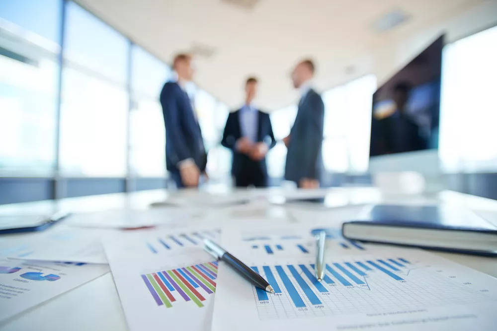 Documents containing graphs lay on a desk in the foreground with three professional speaking to each other in the background.