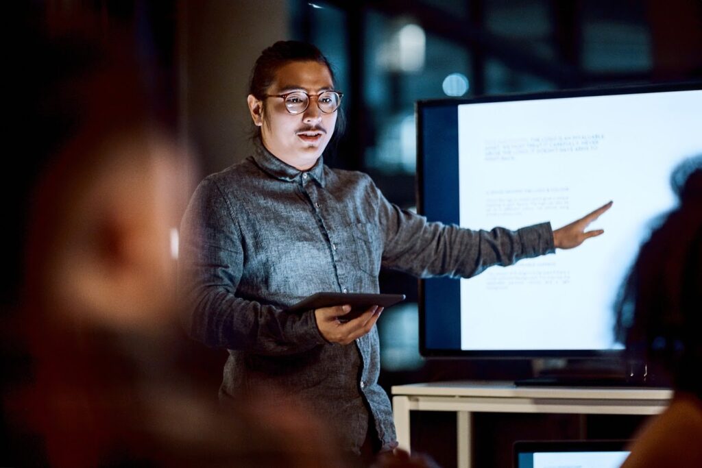 A data analyst holds a tablet and gestures to a monitor during a presentation.